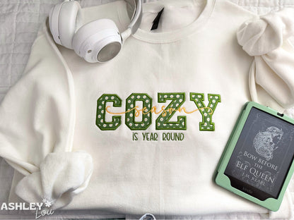 White sweatshirt with 'COZY is year round' text, white headphones, and a green book on a light background.